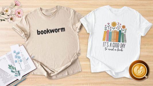 Two cozy bookish t-shirts laid flat on a wooden background with an open book and coffee, featuring “bookworm” and “it’s a good day to read a book” designs