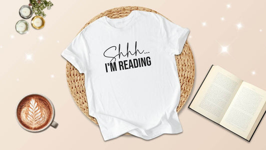 Bookish aesthetic flatlay with a ‘Shhh… I’m reading’ T-shirt, open book, latte art coffee and candles — perfect gift idea for bookish, romantasy and dark romance readers.